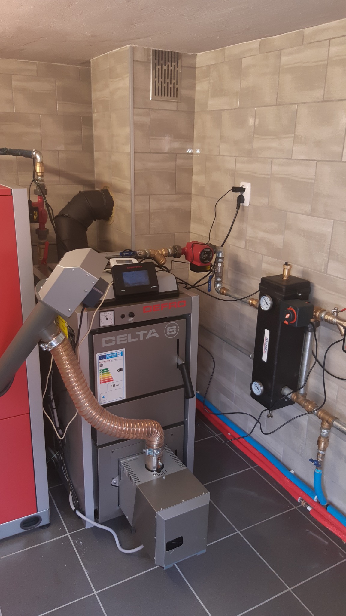 Szary piec na paliwo stałe Delta S z podajnikiem, widoczne rury instalacji CO w kolorze czerwonym i niebieskim, na tle wykafelkowanej ściany