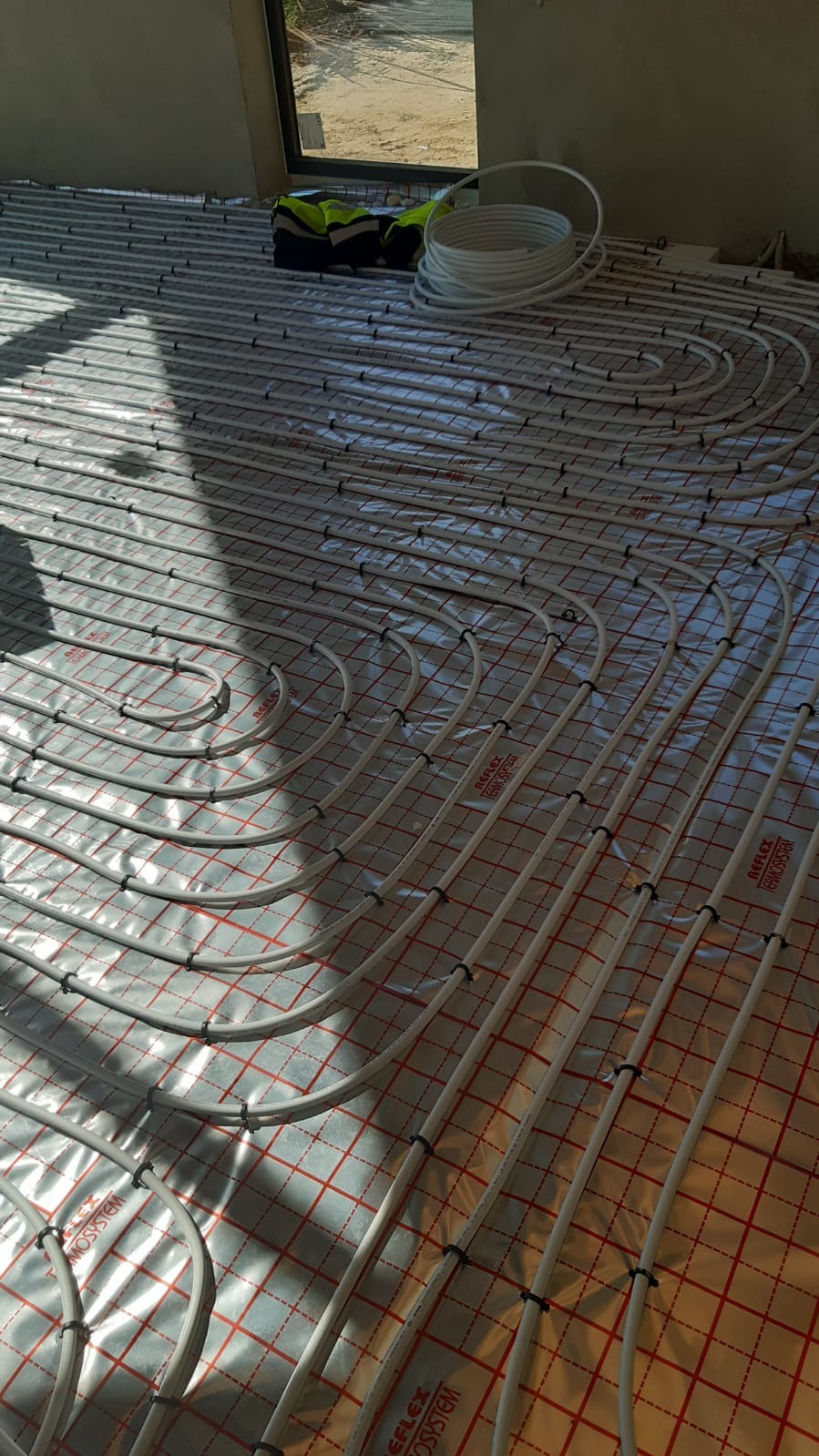 Układanie rur ogrzewania podłogowego na izolacji z czerwoną siatką, widoczny fragment zwiniętej rury i odblaskowa kamizelka przy oknie.