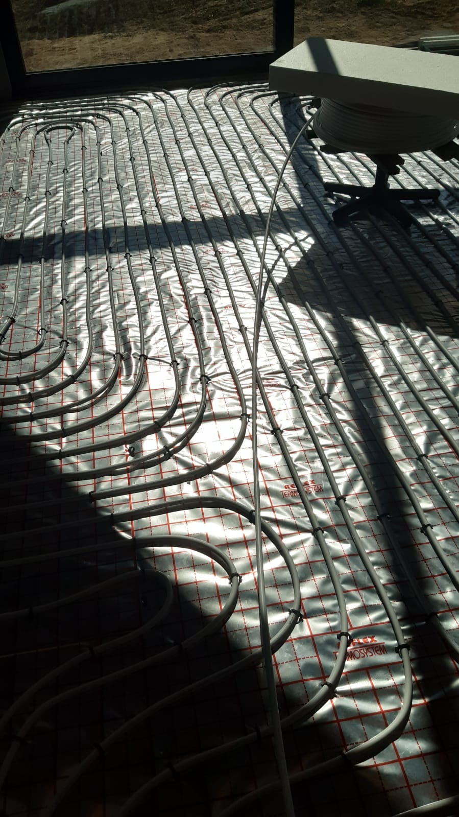 Montaż ogrzewania podłogowego: rury rozprowadzone na srebrnej folii z czerwoną kratką, częściowo w cieniu, z widoczną rolką rury na krześle w rogu.