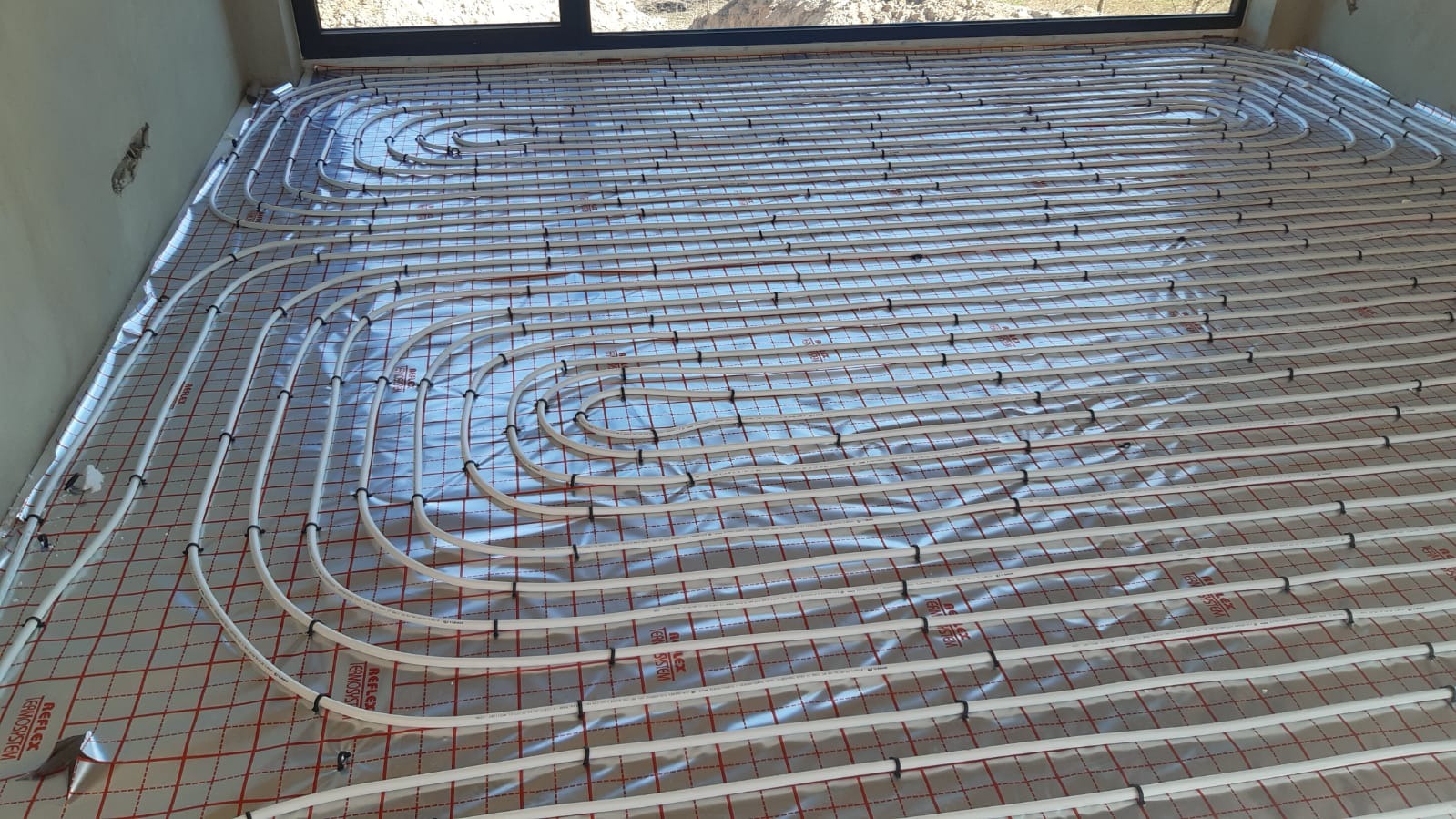 Białe rury ogrzewania podłogowego ułożone na srebrnej folii z czerwoną kratką, widok z góry w jasnym pomieszczeniu z oknem.