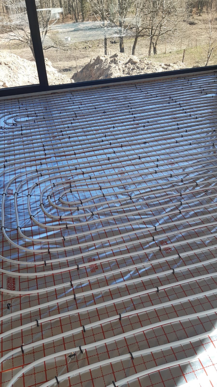 Instalacja ogrzewania podłogowego w Strzelnie: białe rury ułożone na srebrnej folii z czerwoną kratką, widok z góry na przygotowany system przed wylewką.