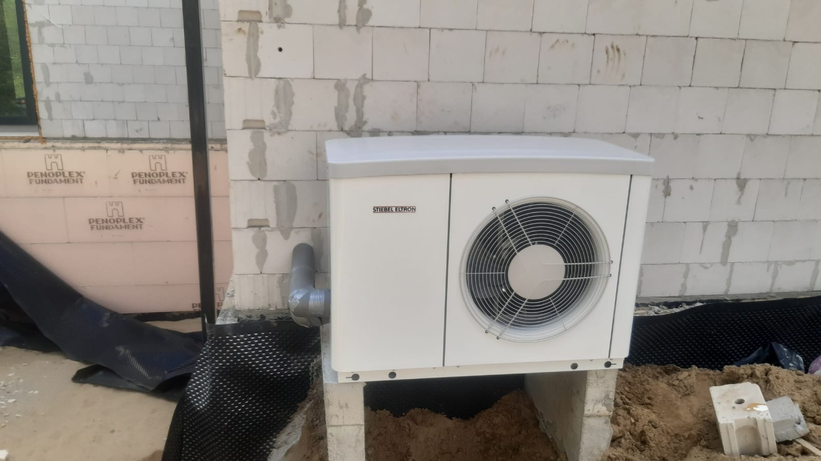 Biała jednostka zewnętrzna z logiem Stiebel Eltron, zamontowana na betonowych wspornikach przy ścianie budynku w trakcie budowy, widoczne ocieplenie fundamentów i fragment okna.