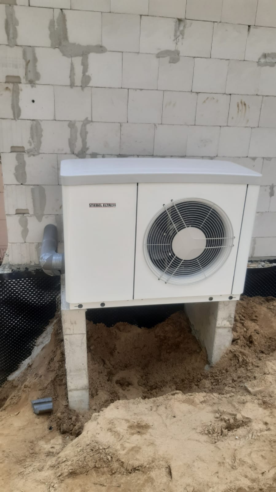 Biała pompa ciepła marki Stiebel Eltron zamontowana na betonowym fundamencie przy ścianie z bloczków betonowych w trakcie budowy, widoczny fragment rury odprowadzającej skropliny.
