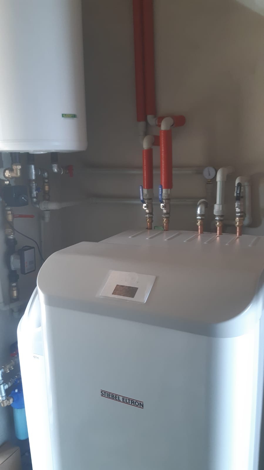 Biały zasobnik ciepłej wody użytkowej marki Stiebel Eltron z podłączonymi rurami i zaworami, widoczne elementy instalacji hydraulicznej w pomieszczeniu technicznym.