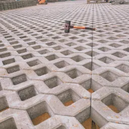 Szara ażurowa nawierzchnia z betonu z widocznym gumowym młotkiem i stosem kostki w tle.