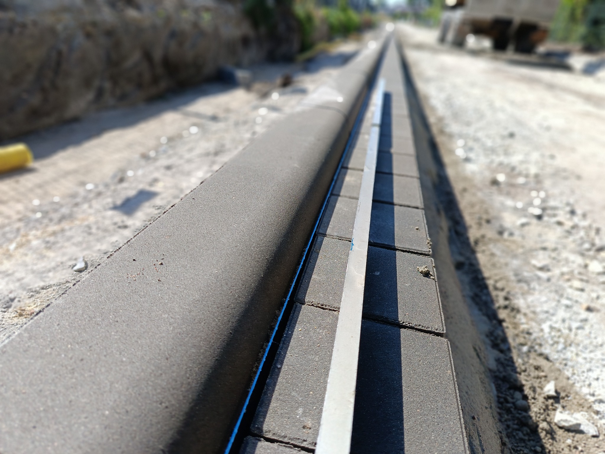 Wyrównywanie krawężnika z kostki brukowej za pomocą poziomicy; widoczne niedokończone podłoże i ciężki sprzęt w tle.