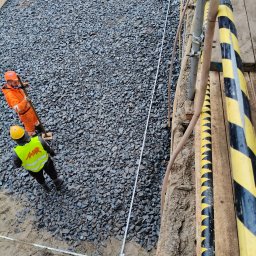 ELWM SPÓŁKA Z OGRANICZONĄ ODPOWIEDZIALNOŚCIĄ - Dwaj pracownicy w odblaskowych ubraniach układają warstwę tłucznia w wykopie pod fundamenty, wyznaczając granice taśmą mierniczą, widok z góry.