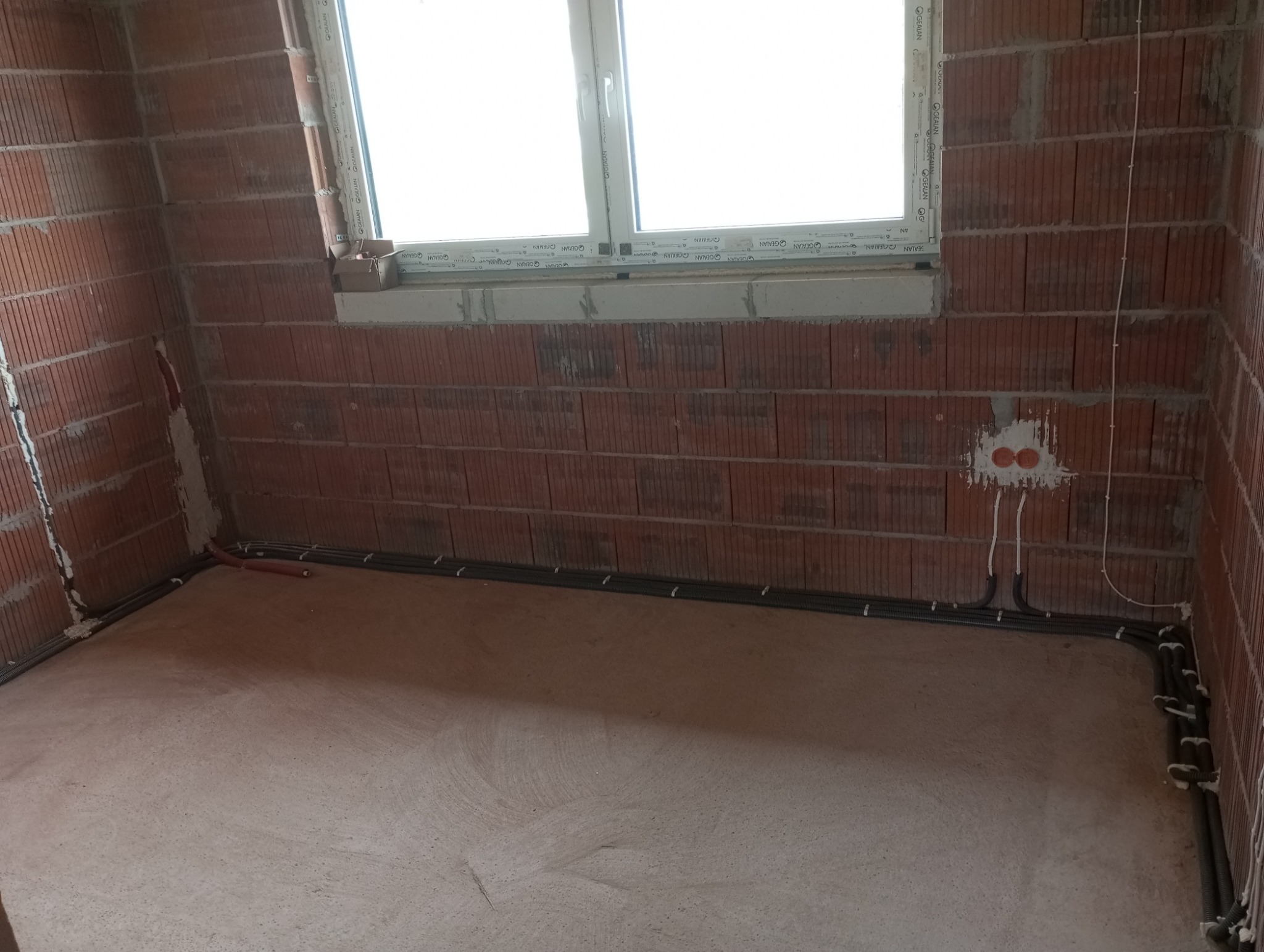 Surowy stan instalacji elektrycznej w nowym domu: widoczne pomarańczowe puszki instalacyjne w ścianie z czerwonej cegły, szare rurki karbowane prowadzące kable po podłodze i ścianach, okno w tle.