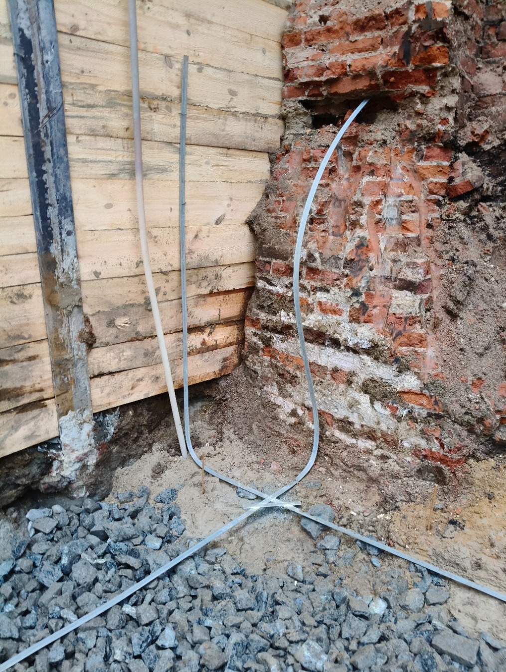 Kabel elektryczny ułożony na gruzie i piasku przy fundamentach budynku z drewnianą szalunką i fragmentem ceglanego muru.