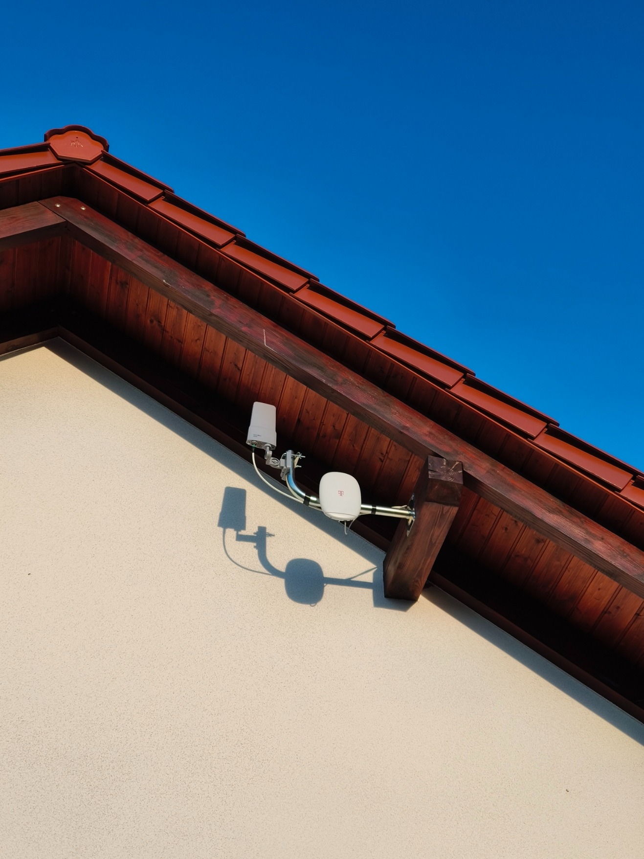 Antena operatora telefonii komórkowej zamontowana na elewacji domu pod okapem dachu z brązową dachówką, widok z dołu w słoneczny dzień.