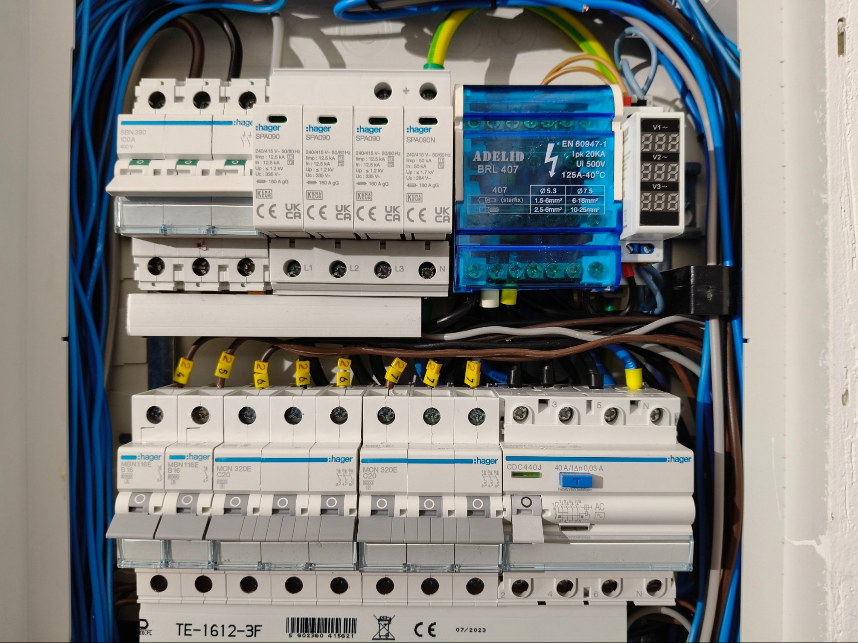 Rozdzielnica elektryczna z zamontowanymi zabezpieczeniami przeciwprzepięciowymi Hager, modułem ADELID BRL 407 i miernikiem napięcia, widoczne kable w kolorach niebieskim, brązowym i żółto-zielonym.