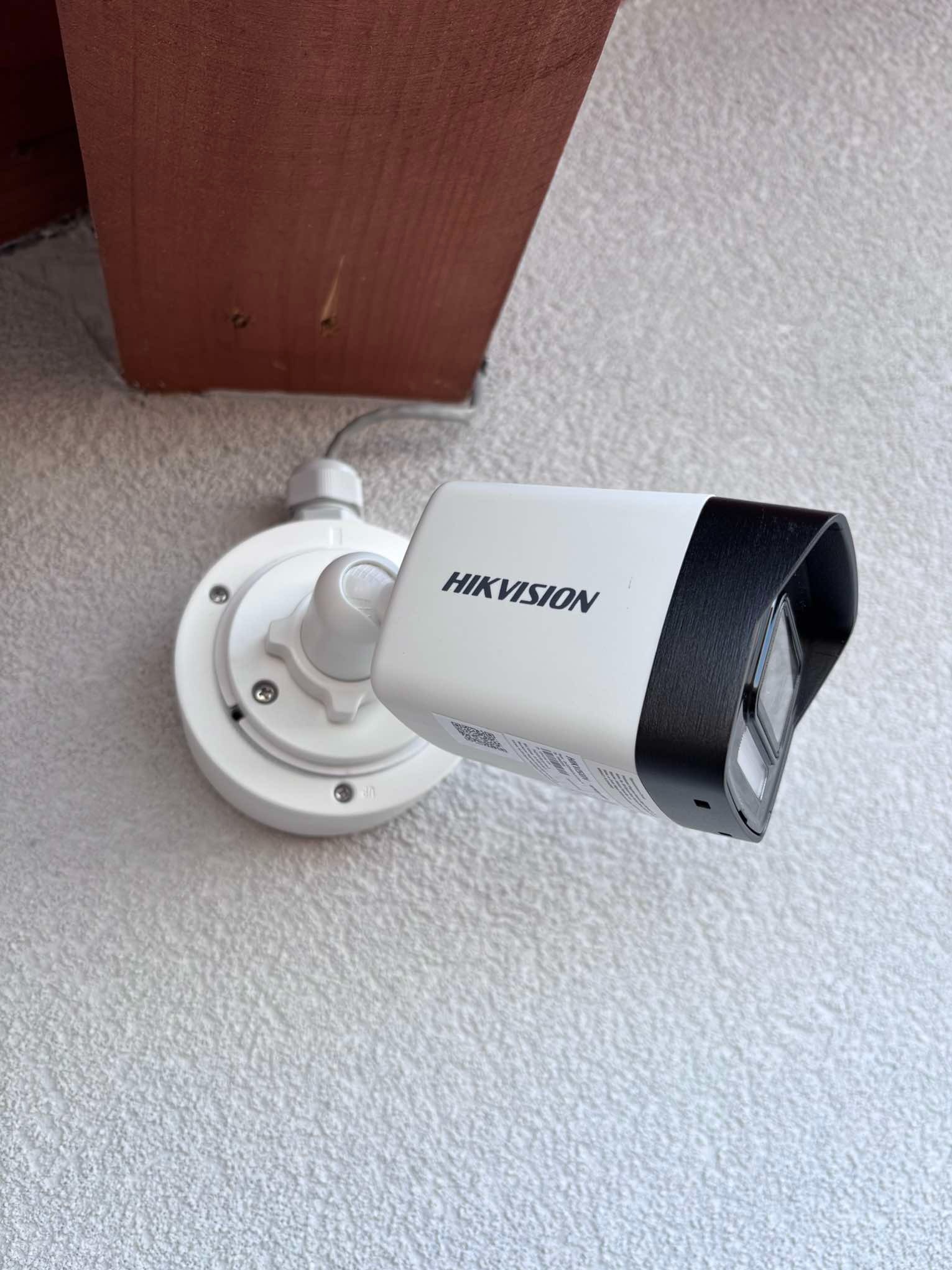 Zewnętrzna kamera monitoringu Hikvision zamontowana na chropowatej, jasnej ścianie, z widocznym kablem i fragmentem drewnianej belki.