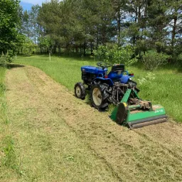 Niebieski traktor z zielonym mulczerem LFN-105 kosi trawę na łące, widoczny pas świeżo skoszonej trawy, w tle drzewa i błękitne niebo.