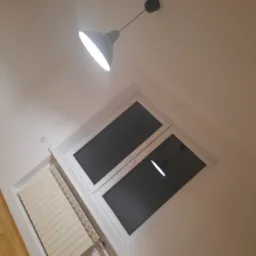 Świeżo pomalowane na biało wnętrze z grzejnikiem i oknem dachowym, oświetlone wiszącą lampą z szarym kloszem.