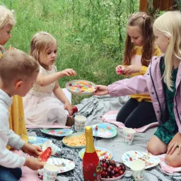Grupa dzieci siedzi na kocu podczas przyjęcia na trawie, częstując się słodyczami z talerzy i popijając napoje z papierowych kubków z napisem 'Happy Birthday'.