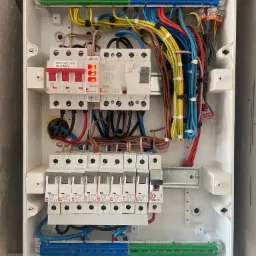 Otwarta biała skrzynka rozdzielcza z bezpiecznikami, przewodami o różnych kolorach i listwami zaciskowymi, widoczne elementy instalacji elektrycznej.