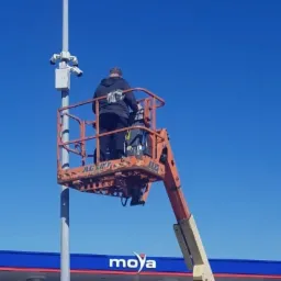 Pracownik na podnośniku koszowym montuje kamery monitoringu na słupie, stacja paliw Moya w tle.