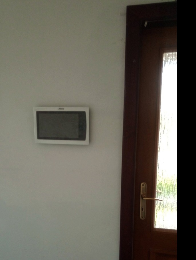 Biały panel sterowania, prawdopodobnie domofonu lub alarmu, zamontowany na jasnej ścianie obok ciemnobrązowych drzwi z ozdobnym szkłem.