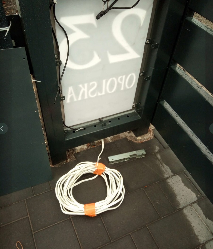 Otwarta obudowa podświetlanego szyldu z numerem posesji '53' i napisem 'OPOLSKA', zwój kabla elektrycznego na szarym bruku, widoczne elementy montażowe i fragment ogrodzenia.