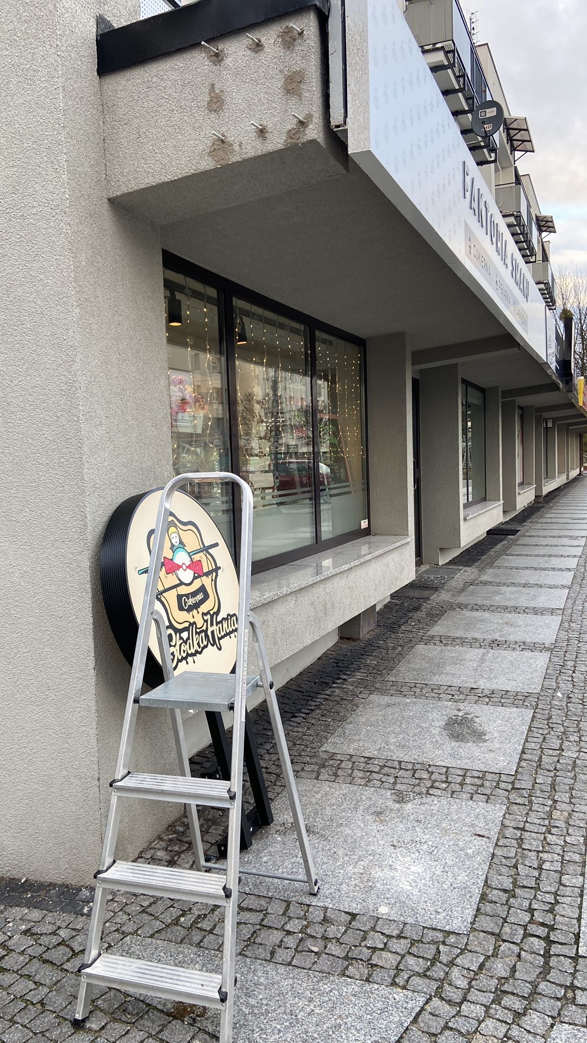 Aluminiowa drabina oparta o ścianę budynku obok okrągłego szyldu 'Słodka Hania' na tle chodnika z kostki brukowej i fasady budynku z oknami.