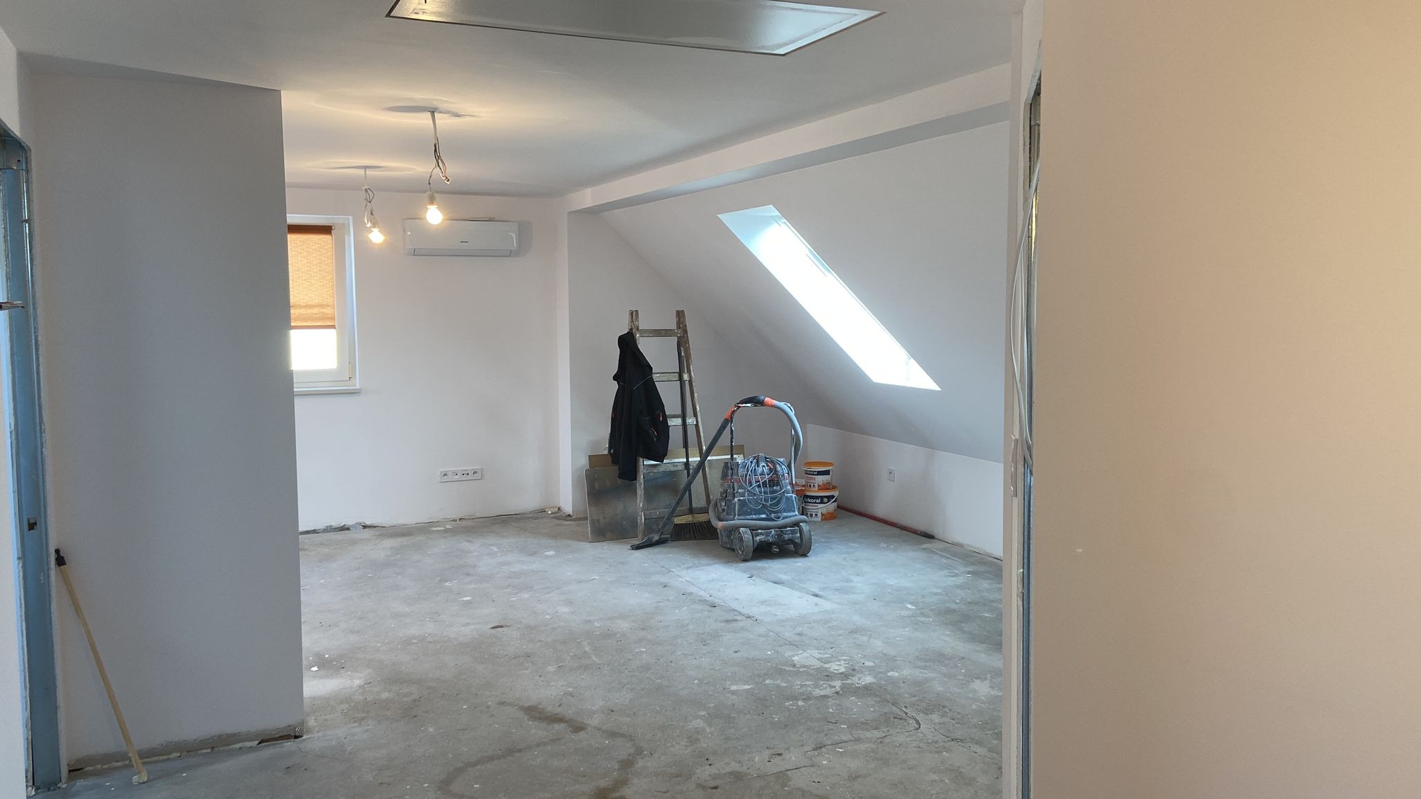 Poddasze w trakcie remontu: szlifierka do betonu, drabina oparta o ścianę, okno dachowe w skosie, ślady szlifowania na podłodze, wiszące żarówki bez kloszy, klimatyzator na ścianie, widoczne puszki...