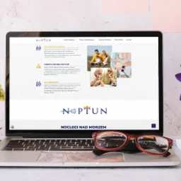 Laptop wyświetlający stronę internetową z ofertą noclegów nad morzem i turnusów rehabilitacyjnych, z okularami na klawiaturze i kwiatami w wazonach po bokach.