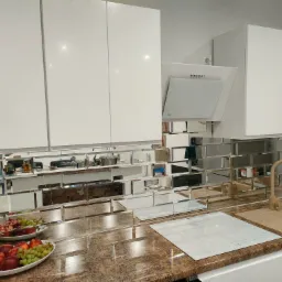 Kuchnia z białymi szafkami, szklanym panelem ściennym w formie małych płytek lustrzanych, brązowym blatem, brązowym zlewem i owocami w misce.