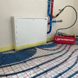 Instalacja ogrzewania podłogowego z niebieskich rur na folii z czerwoną kratką, widoczna skrzynka rozdzielcza i manometr Rothenberger.