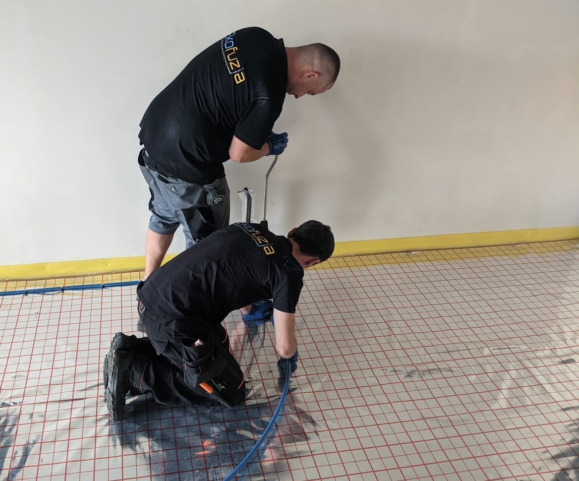 Dwóch instalatorów w czarnych koszulkach z logo firmy montuje ogrzewanie podłogowe na folii z siatką, jeden stoi, drugi klęczy, używając narzędzi.