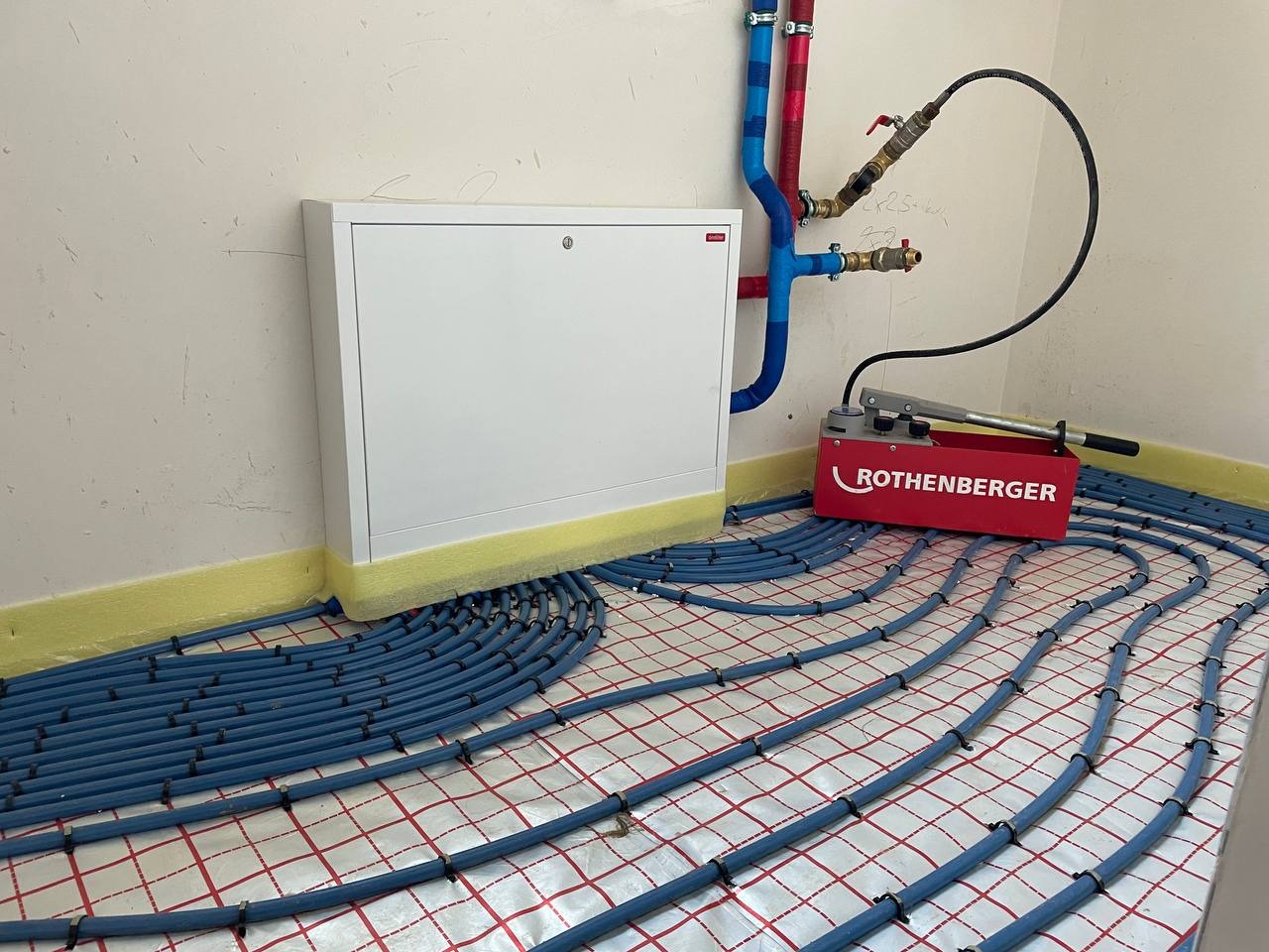 Instalacja ogrzewania podłogowego z niebieskich rur na folii z czerwoną kratką, widoczna skrzynka rozdzielcza i manometr Rothenberger.