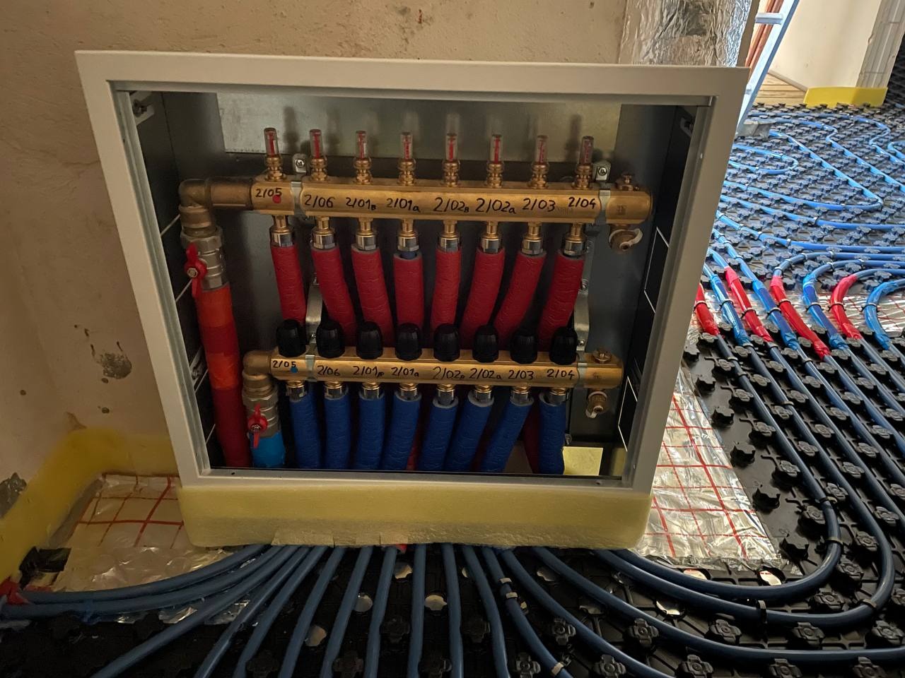 Rozdzielacz ogrzewania podłogowego w obudowie, widoczne rury doprowadzające i odprowadzające wodę w kolorach czerwonym i niebieskim, instalacja w trakcie montażu.