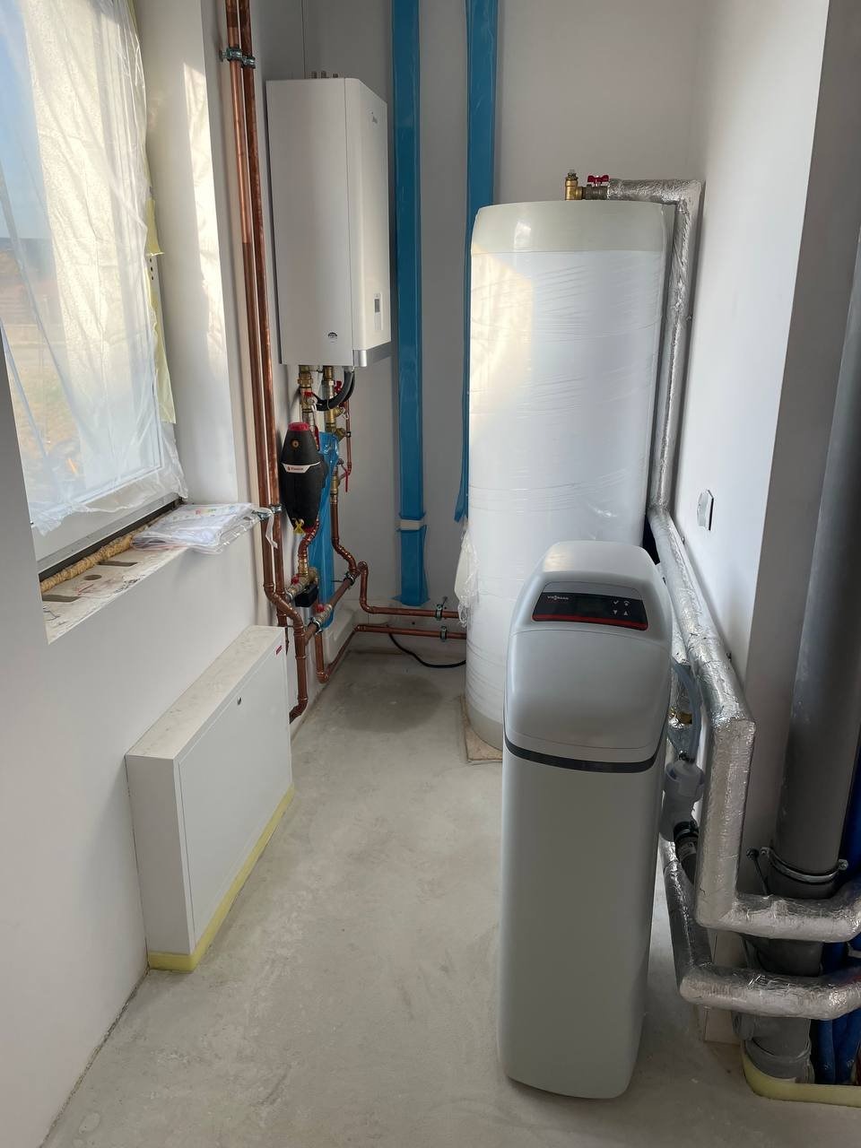 Kotłownia w trakcie montażu: biały piec gazowy, zasobnik ciepłej wody owinięty folią, zmiękczacz wody Viessmann oraz rury miedziane i niebieskie PCV.