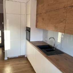 Nowoczesna kuchnia w Gdańsku: białe, minimalistyczne szafki dolne i wysokie zabudowy, górne szafki z drewnopodobnym fornirem, czarny zlew podwieszany, bateria chromowana, drewniana podłoga.