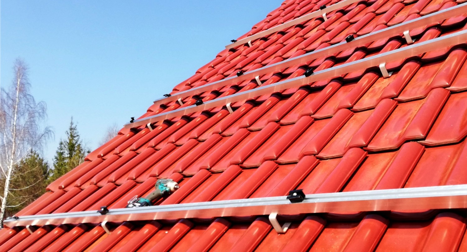 Montaż szyn pod panele słoneczne na czerwonym dachu z dachówki ceramicznej, wiertarko-wkrętarka leży na szynie, widoczne uchwyty mocujące i błękitne niebo w tle.