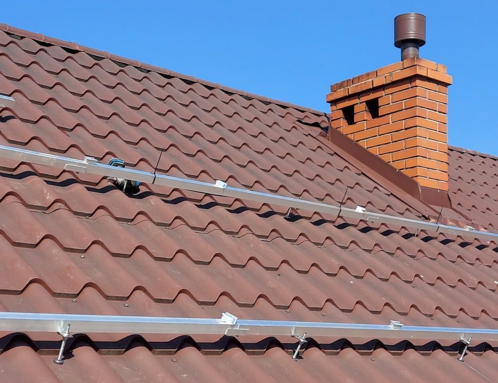 Montaż szyn pod panele fotowoltaiczne na dachu z brązowej dachówki ceramicznej, widoczny komin z cegły i fragment wiertarki akumulatorowej.