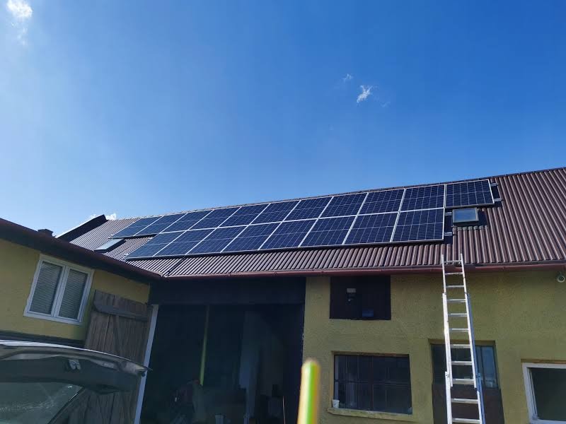 Instalacja paneli słonecznych na falistej, brązowej blasze dachu budynku gospodarczego; drabina oparta o ścianę.