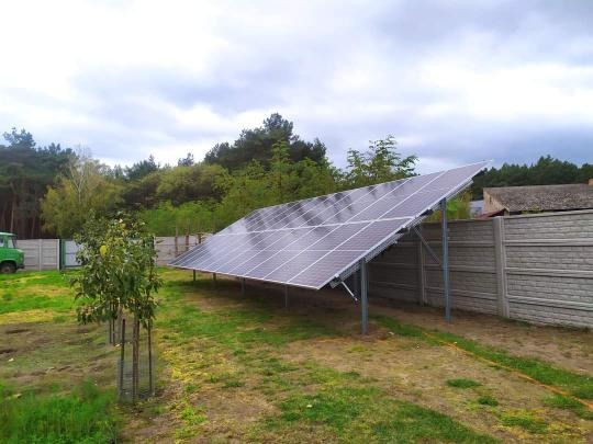 Panel słoneczny umieszczony na metalowej konstrukcji w ogrodzie, widoczny betonowy płot i drzewa w tle, pochmurne niebo.