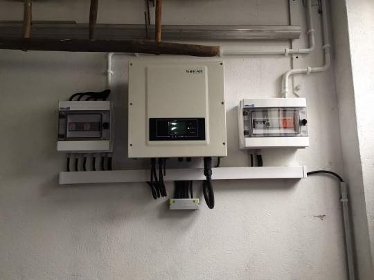 Biały inwerter z podłączonymi kablami i dwoma skrzynkami bezpiecznikowymi na białej ścianie, widoczne elementy instalacji elektrycznej i drabina w górnej części kadru.