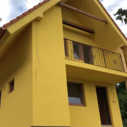 Żółty dom z balkonem, brązową stolarką okienną i dachem z czerwonej dachówki, widoczny z dołu pod kątem, z elementami drewnianymi podtrzymującymi balkon.