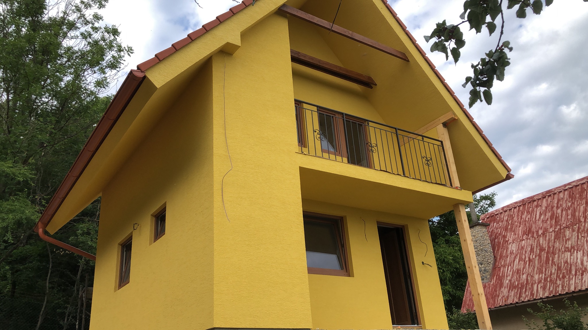 Żółty dom z balkonem, brązową stolarką okienną i dachem z czerwonej dachówki, widoczny z dołu pod kątem, z elementami drewnianymi podtrzymującymi balkon.