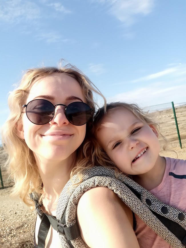 Uśmiechnięta kobieta w okularach przeciwsłonecznych robiąca selfie z małą dziewczynką w nosidełku na tle błękitnego nieba i piaszczystego terenu.