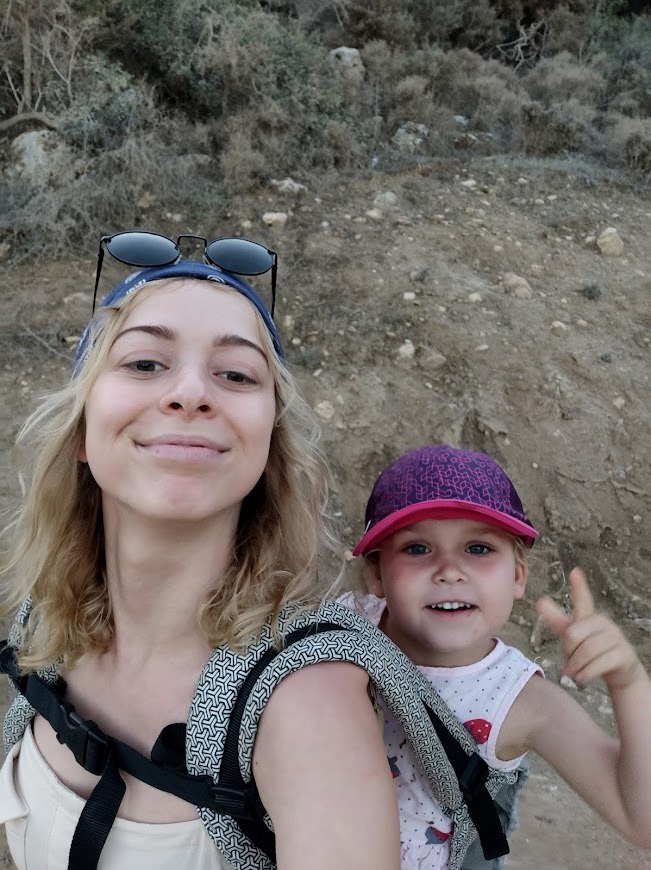 Kobieta z blond włosami w chuście na głowie, niosąca w nosidle małą dziewczynkę w różowej czapce, uśmiechnięte, na tle skalistego wzgórza w okolicach Krakowa.