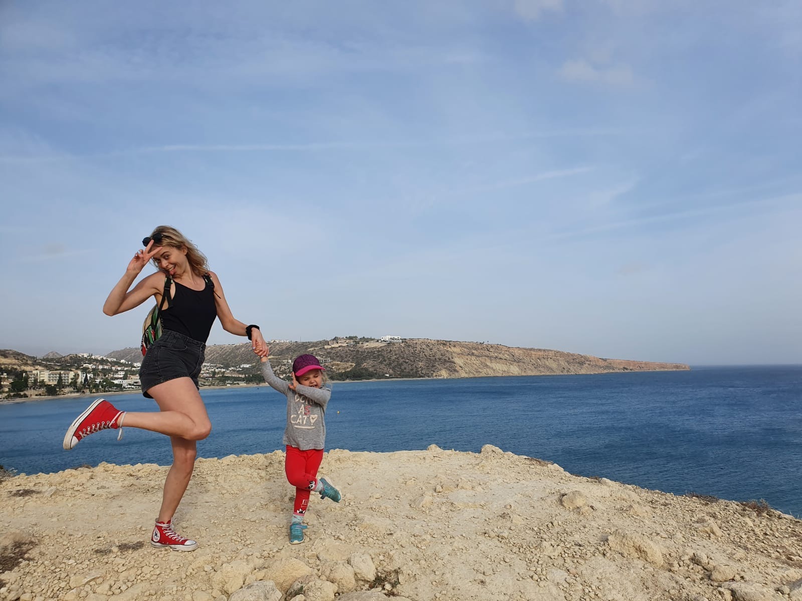 Uśmiechnięta kobieta w czerwonych trampkach i córka w czapce z daszkiem trzymają się za ręce na tle morskiego krajobrazu podczas spaceru.