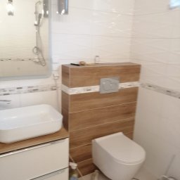 SOLID-BUD - Nowoczesna łazienka z podwieszaną toaletą, umywalką nablatową na szafce z szufladami i lustrem, wykończona białymi i drewnopodobnymi płytkami, z widocznymi akcesoriami do sprzątania.