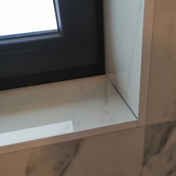 SOLID-BUD - Fragment okna z czarną ramą i marmurowym parapetem, wykończonymi płytkami imitującymi marmur na ścianie.
