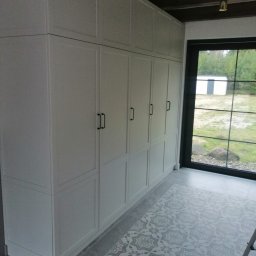 SOLID-BUD - Wysoka, zabudowana szafa wnękowa z szarymi frontami i czarnymi, minimalistycznymi uchwytami, na tle okna z widokiem na zieleń, z ozdobną, wzorzystą podłogą z płytek w odcieniach szarości.