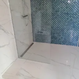 Wykończona kabina prysznicowa z niebieską mozaiką w kształcie marokańskiej koniczyny na ścianie, białe marmurowe płytki na pozostałych ścianach i podłodze, odpływ liniowy przy szklanej ściance.