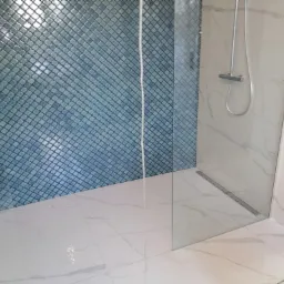 Wykończona kabina prysznicowa z niebieską mozaiką na ścianie i białymi płytkami imitującymi marmur na podłodze i pozostałych ścianach, widoczny odpływ liniowy.