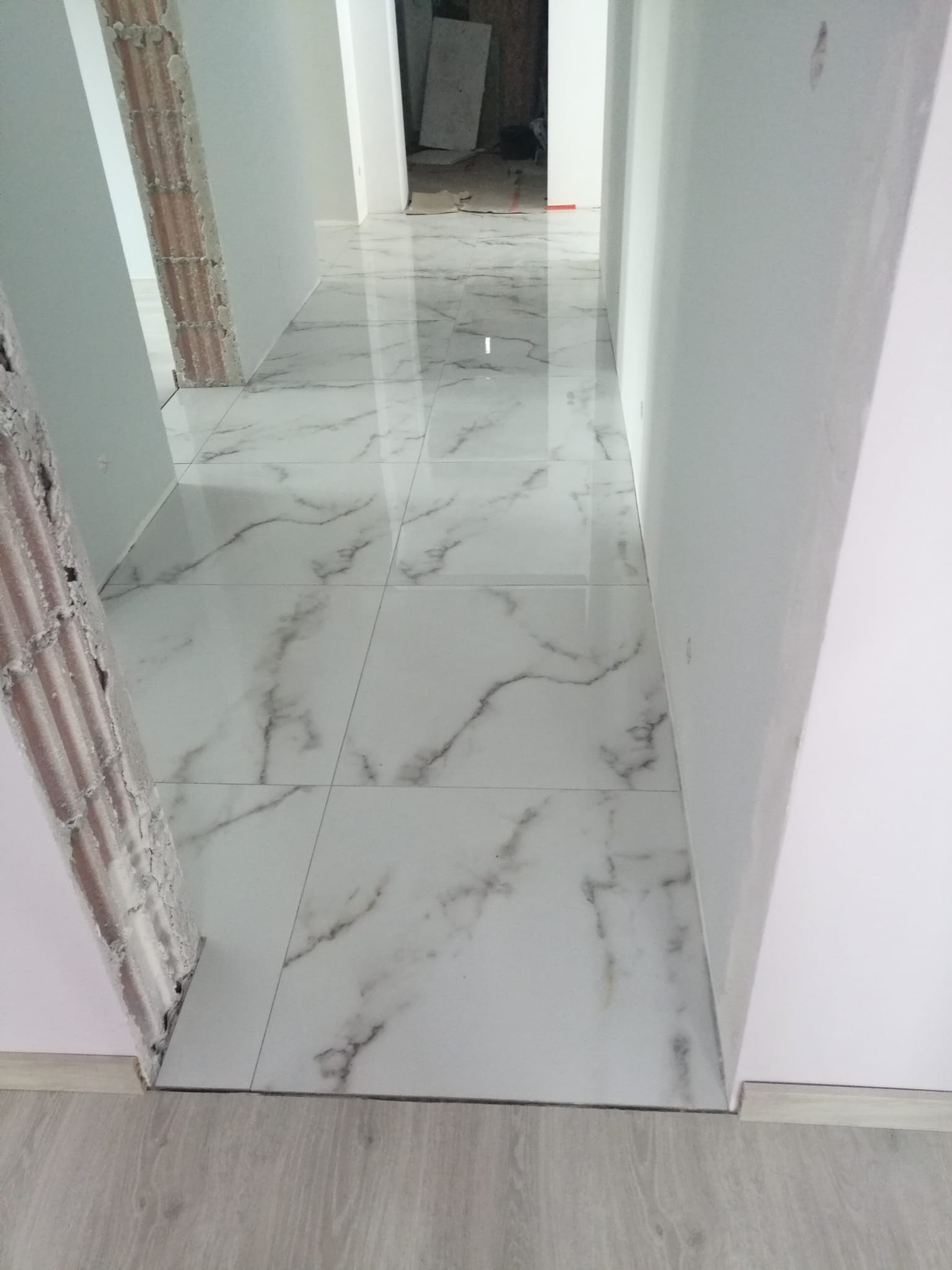 Położona, błyszcząca, biała podłoga z wzorem marmuru w wąskim korytarzu, widoczne surowe wykończenia ścian i podłogi w sąsiednim pomieszczeniu.