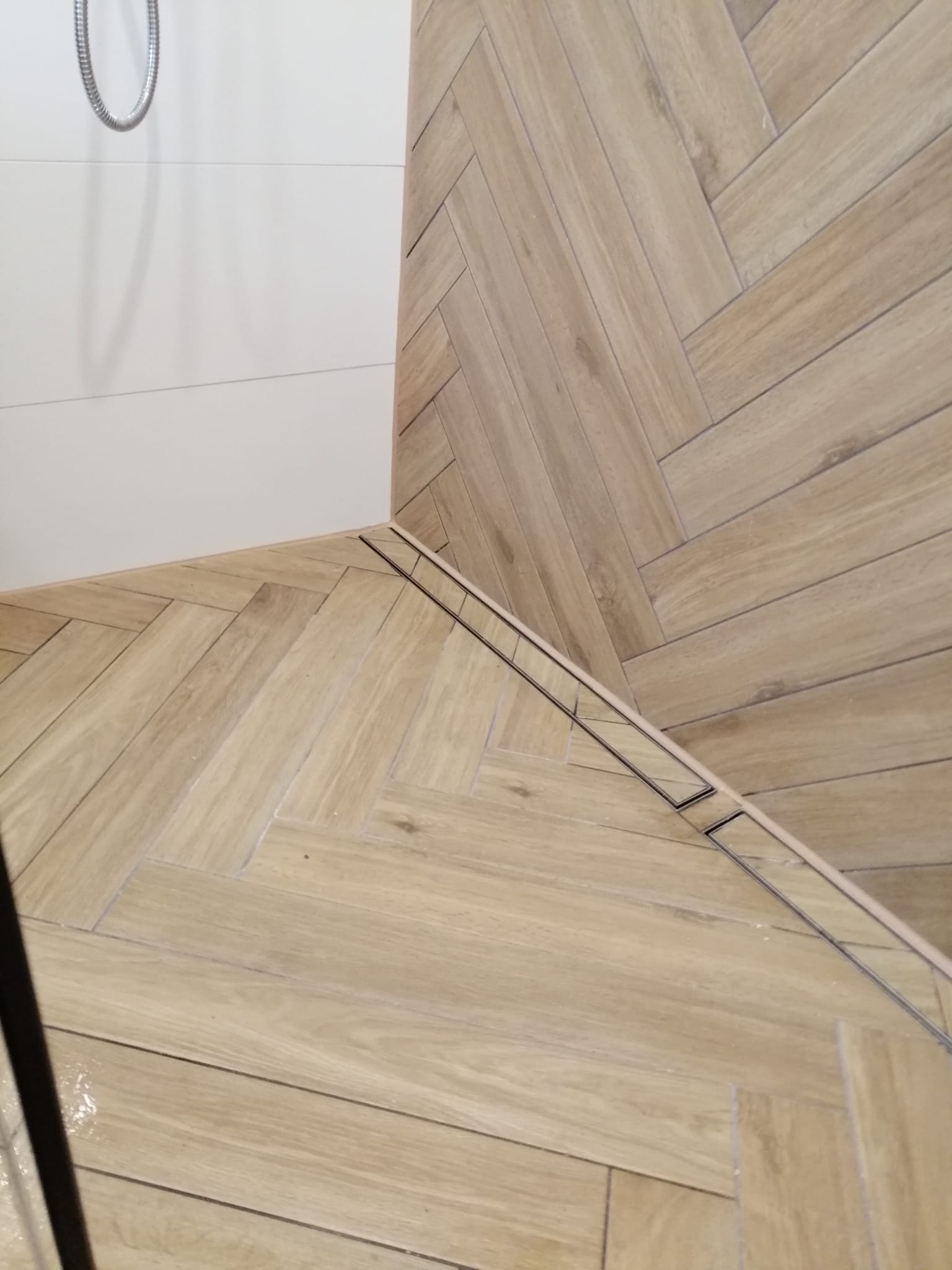 Ułożona w jodełkę podłoga z płytek imitujących drewno w jasnym odcieniu, z widocznym odpływem liniowym pod prysznicem i fragmentem białej ściany wykończonej płytkami.